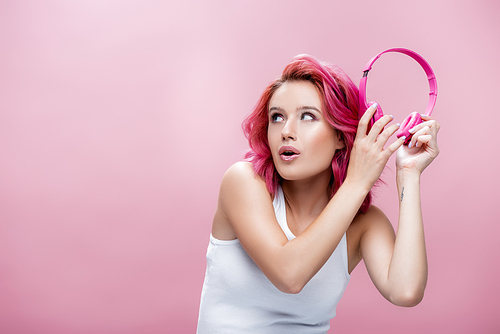 surprised young woman with colorful hair holding headphones near ear isolated on pink