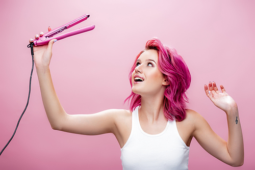 young woman with colorful hair looking at straightener isolated on pink