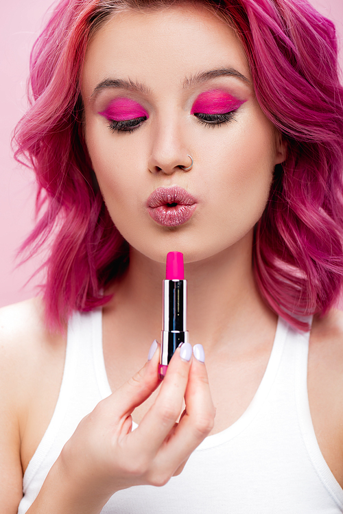 young woman with colorful hair holding lipstick and pouting lips isolated on pink