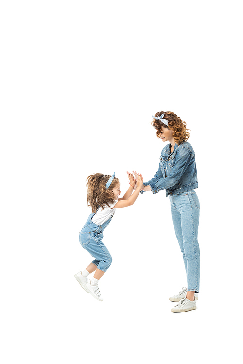 side view of daughter in denim outfit jumping and clapping hands with mother isolated on white