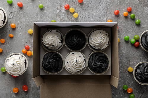 top view of delicious Halloween cupcakes in box near scattered colorful bonbons on concrete grey surface