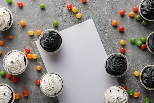 top view of delicious Halloween cupcakes with scattered colorful bonbons near white blank paper on concrete grey surface