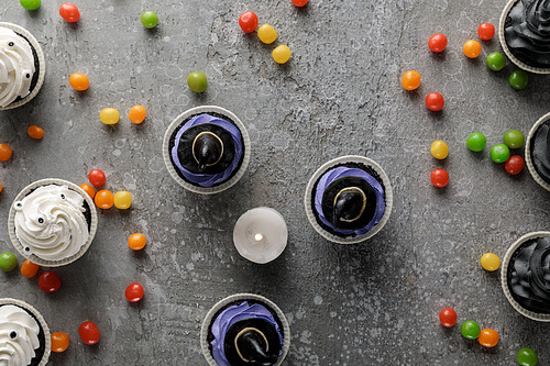 top view of delicious Halloween cupcakes and burning candle with scattered colorful bonbons on concrete grey surface