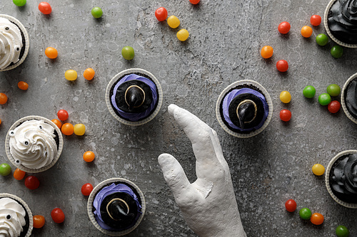 top view of decorative hand near delicious Halloween cupcakes with scattered colorful bonbons on concrete grey surface