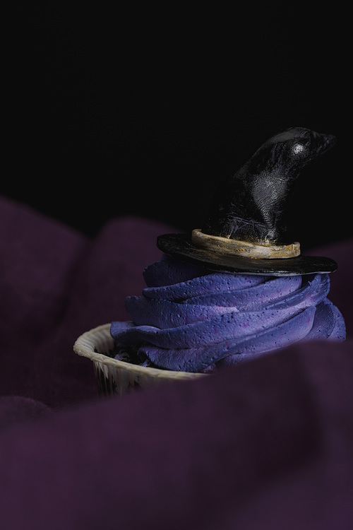 tasty Halloween cupcake with blue cream and decorative witch hat on purple cloth isolated on black