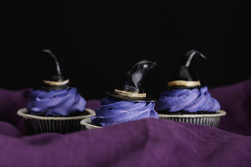 tasty Halloween cupcakes with blue cream and decorative witch hats on purple cloth isolated on black