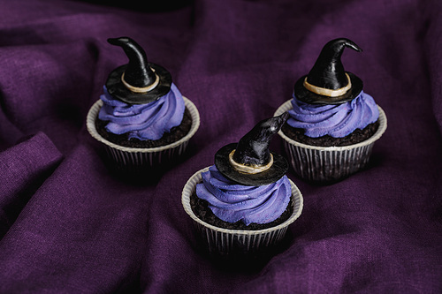 tasty Halloween cupcakes with blue cream and decorative witch hats on purple cloth