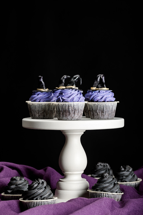tasty Halloween cupcakes on white stand on purple cloth isolated on black