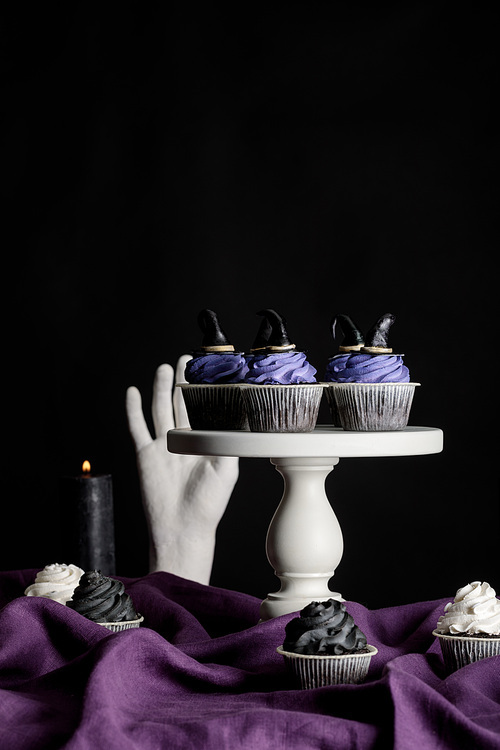 tasty Halloween cupcakes on white stand near burning candles and decorative hand on purple cloth isolated on black