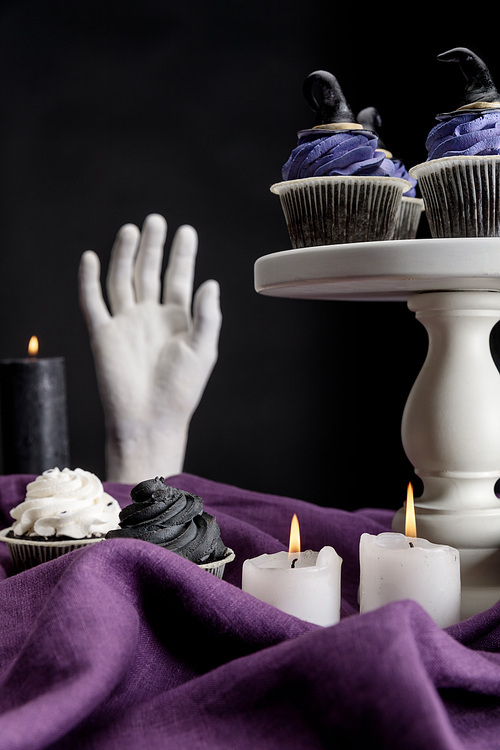 tasty Halloween cupcakes on white stand near burning candles and decorative hand on purple cloth isolated on black