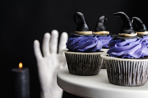 selective focus of tasty Halloween cupcakes with blue cream and decorative witch hats on white stand isolated on black