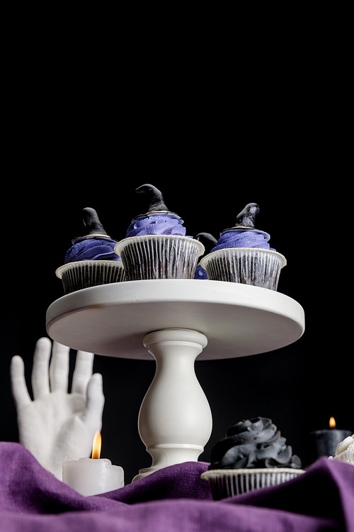 low angle view of tasty Halloween cupcakes on white stand near burning candles on purple cloth isolated on black
