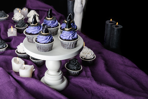tasty Halloween cupcakes on white stand near burning candles on purple cloth isolated on black