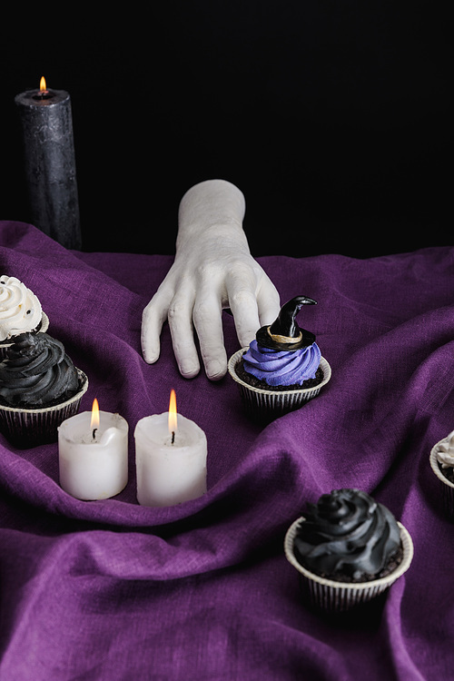 decorative hand near tasty Halloween cupcakes and burning candles on purple cloth isolated on black