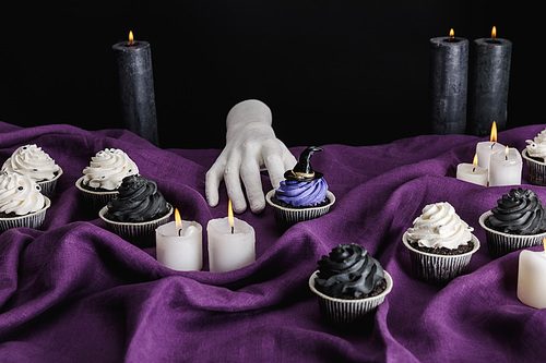 decorative hand near tasty Halloween cupcakes and burning candles on purple cloth isolated on black