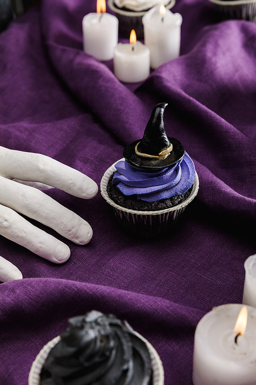 decorative hand near tasty Halloween cupcake with blue cream and decorative witch hat near burning candles on purple cloth