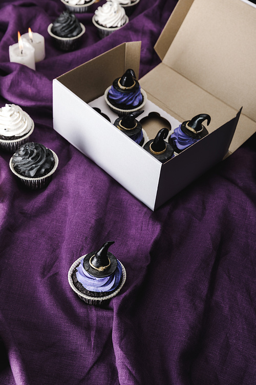 tasty Halloween cupcakes with blue cream and decorative witch hats in box near burning candles on purple cloth