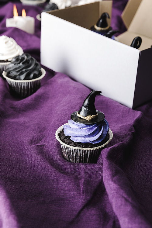 tasty Halloween cupcake with blue cream and decorative witch hat on purple cloth