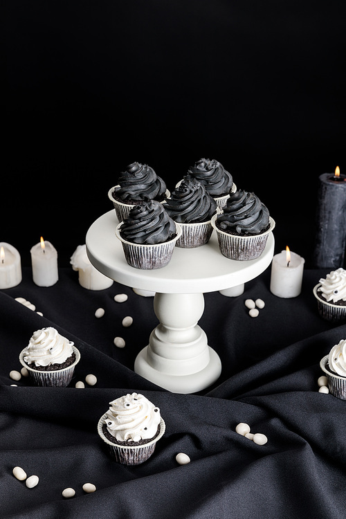 tasty Halloween cupcakes with black cream on white stand near burning candles isolated on black