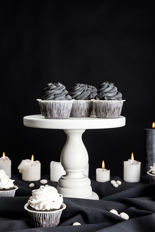 tasty Halloween cupcakes with black cream on white stand near burning candles isolated on black