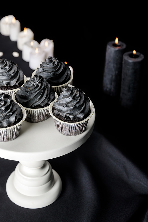 selective focus of tasty Halloween cupcakes with black cream on stand near burning candles on black background