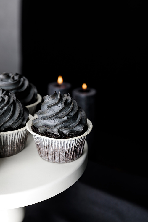 selective focus of tasty Halloween cupcakes with black cream on stand near burning candles isolated on black