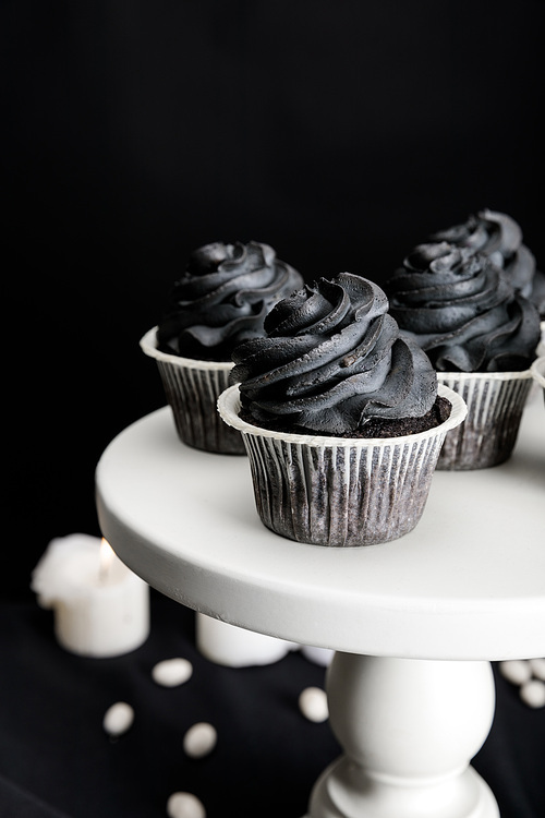 tasty Halloween cupcakes with black cream on stand near burning candles isolated on black