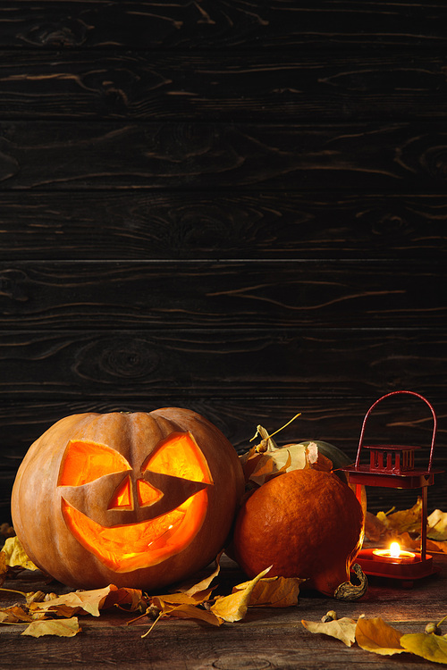 carved spooky Halloween pumpkin, autumnal leaves and burning candle on wooden rustic table on black background