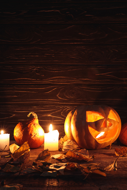 carved spooky Halloween pumpkin, autumnal leaves and burning candles on wooden rustic table