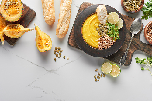 top view of autumnal mashed pumpkin soup on wooden cutting board near ingredients and bread on marble surface