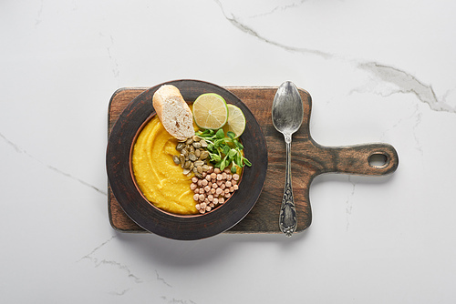 top view of autumnal mashed pumpkin soup in bowl on wooden cutting board with spoon on marble surface