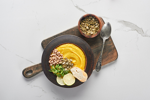 top view of autumnal mashed pumpkin soup on wooden cutting board with seeds and spoon on marble surface