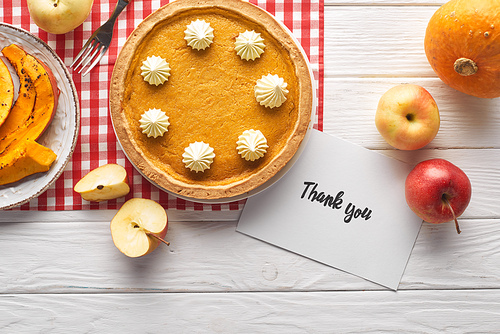 top view of traditional pumpkin pie with thank you card on wooden white table with apples