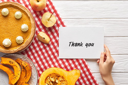 cropped view of woman holding thank you card near pumpkin pie on wooden white table with apples