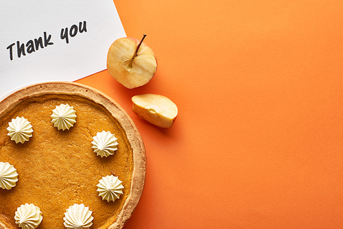 top view of pumpkin pie with thank you card on orange background with apples