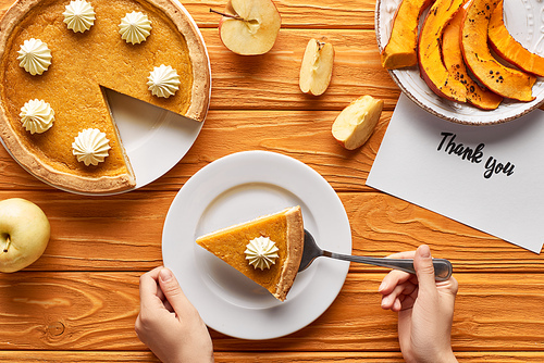 cropped view of woman putting piece of pumpkin cake on plate near thank you card on wooden table with apples