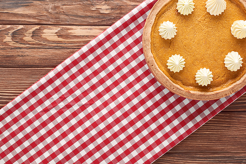 top view of delicious pumpkin pie with whipped cream on checkered napkin on brown wooden surface