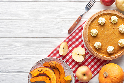 tasty pumpkin pie with whipped cream on checkered napkin near raw and sliced baked pumpkins, cut and whole apples, and fork on white wooden table