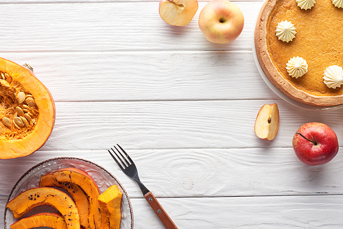 tasty pumpkin pie with whipped cream near raw and sliced baked pumpkin, cut and whole apples, and fork on white wooden table