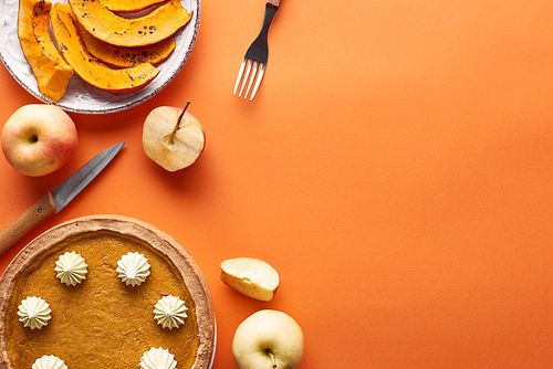 tasty pumpkin pie with whipped cream near sliced baked pumpkin, whole and cut apples, knife and fork on orange surface
