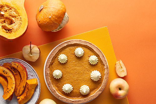 delicious pumpkin pie with whipped cream on textured napkin near raw and baked pumpkins, cut and whole apples on orange surface