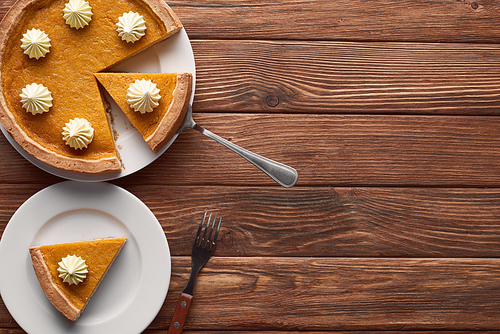 tasty pumpkin pie with whipped cream near spatula and fork on brown wooden surface