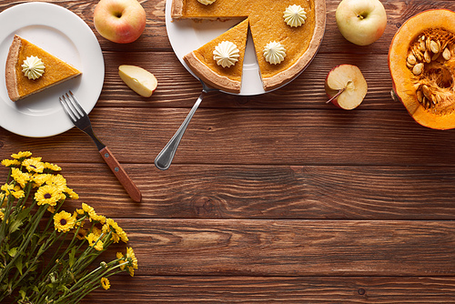 tasty pumpkin pie with whipped cream near spatula and fork, half of raw pumpkin, whole and cut apples on brown wooden surface