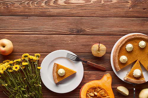 tasty pumpkin pie with whipped cream near fork, half of raw pumpkin, cut and whole apples, and yellow flowers on brown wooden table