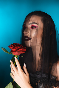 evil bride with black makeup looking away through veil and holding rose on blue