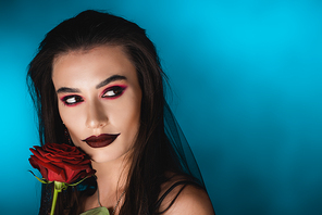 young woman with black makeup in veil looking away near rose on blue