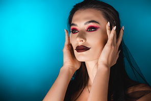 young woman with black makeup touching hair and looking away on blue