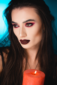 pale woman with makeup and veil looking away while holding burning candle on blue