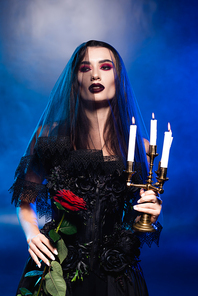 brunette woman in black dress and veil holding red rose and burning candles on blue with smoke, halloween concept