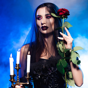 woman in black dress and veil holding red rose and burning candles on blue with smoke, halloween concept